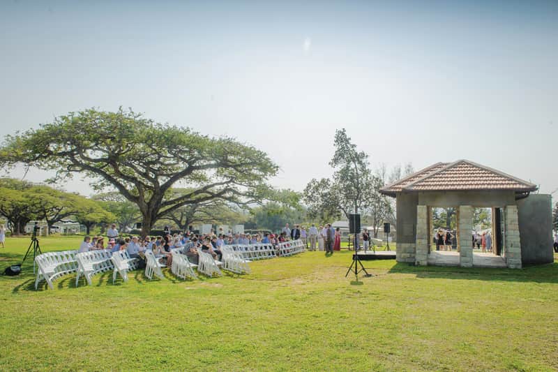 Rows of white chairs on a sunlit lawn where the family gathers for the 125-year dedication service, beside a broad acacia tree and a stone pavilion