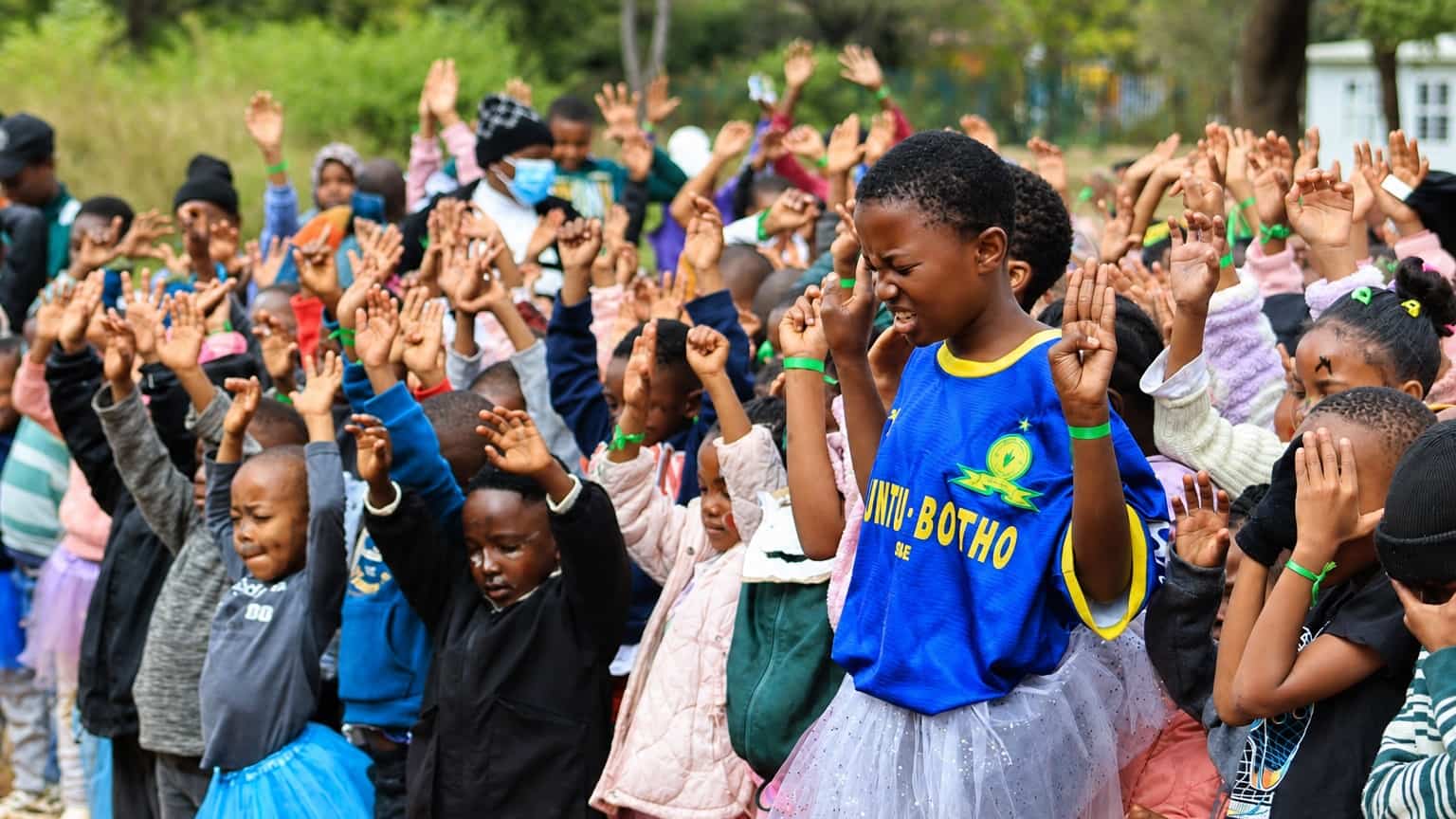 A large group of young children outdoors with their hands raised, a girl in a blue football shirt in the foreground
